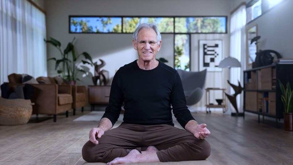 video-thumb – Jon Kabat-Zinn lächelnd in Meditationshaltung in einem lichtdruchfluteten, geschmackvoll eingerichteten Wohnzimmer